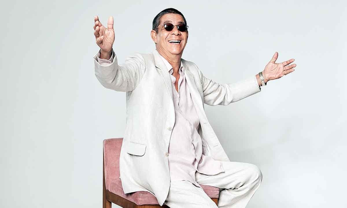 Usando óculos escuros e terno branco, Zeca Pagodinho sorri, com os braços abertos, sentado numa poltrona -  (crédito: Guto Costa/divulgação)