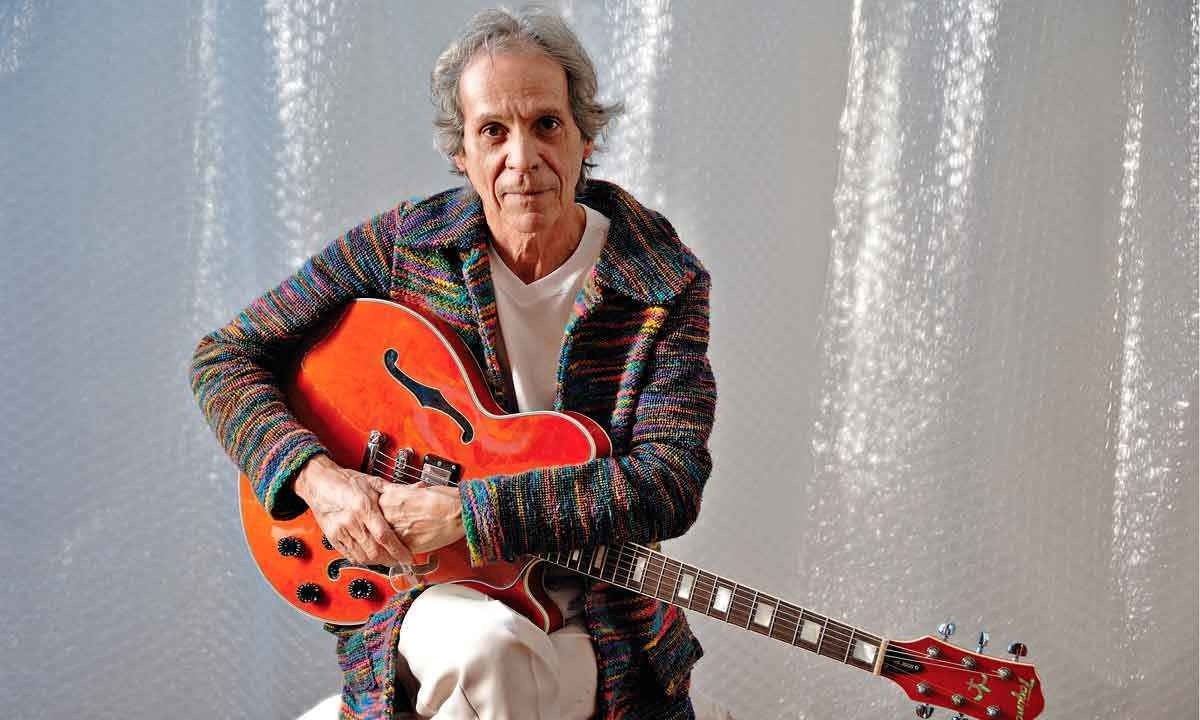 Beto Guedes olha para a câmera, sentado num banco, segurando a guitarra e usando casaco colorido -  (crédito: Cristiano Quintino/divulgação)