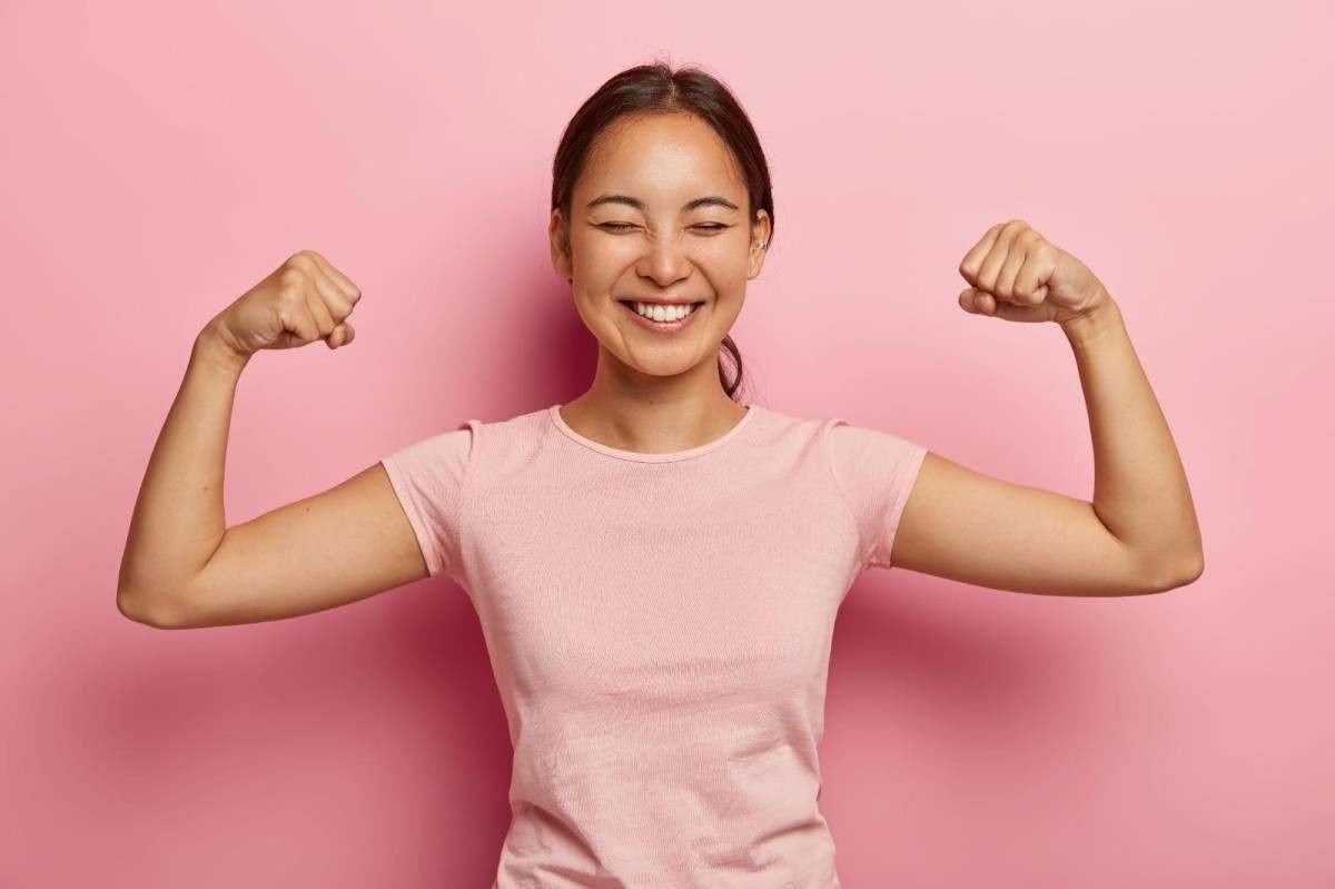 Uma mulher, vestindo blusa rosa de mangas curtas, dobra os braços para cima, demonstrando a força feminina em seguir lutando pela igualdade de direitos 