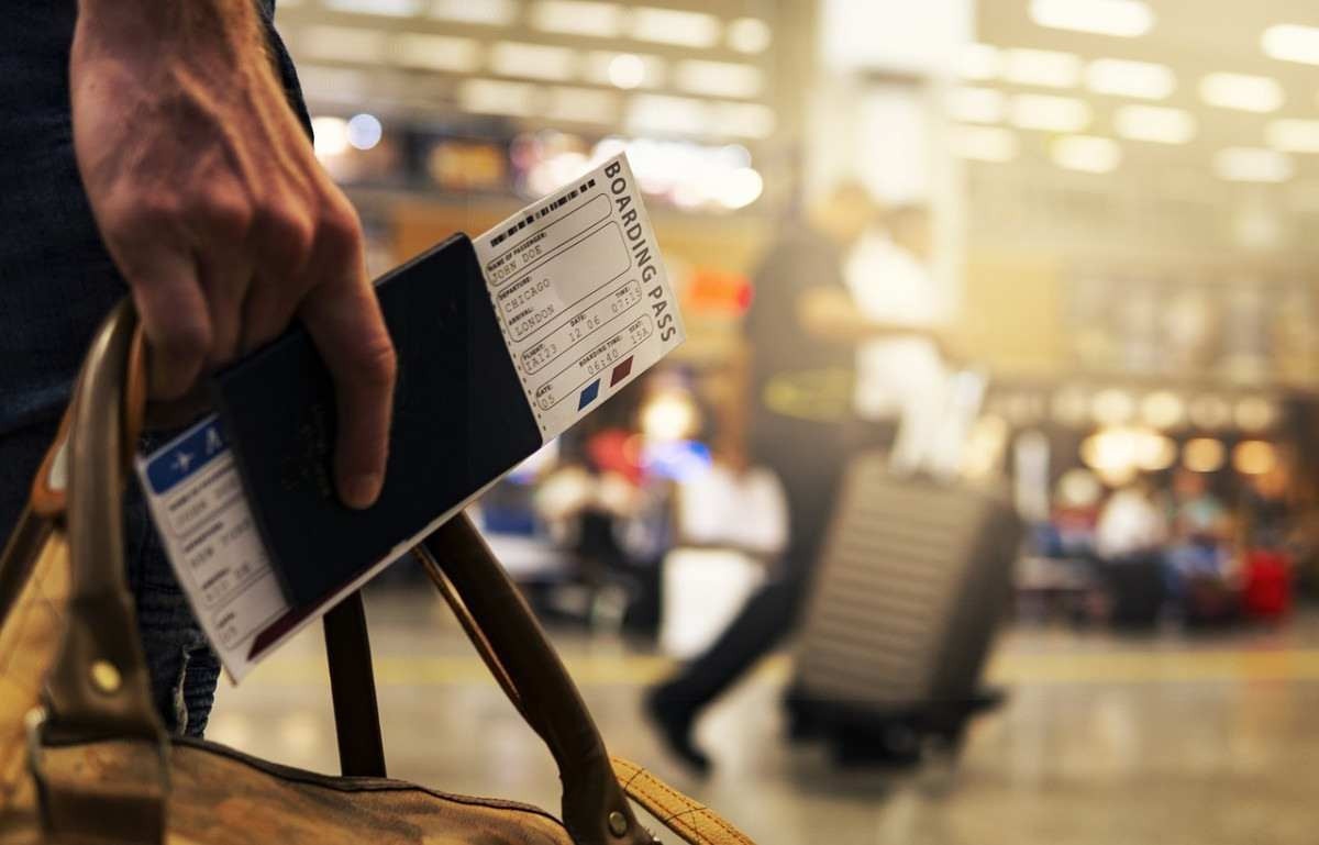 Em primeiro plano, à esquerda, está em destaque uma mão direita segurando um passaporte e uma mala de mão. No segundo plano, o cenário é de um aeroporto.