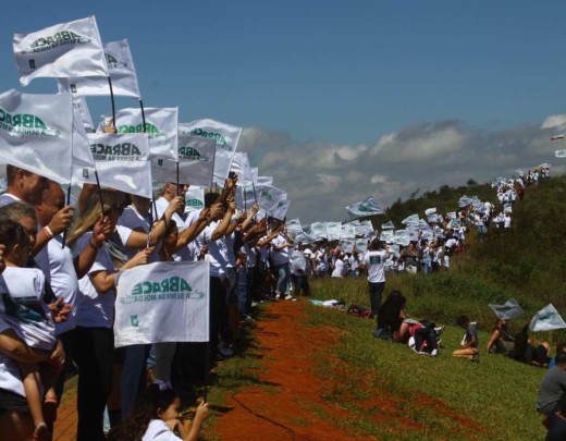 Foto mostra centenas de pessoas lada a lado, como se fosse uma fila, abraçando simbolicamente a Serra da Moeda. As pessoas seguram bandeirinhas brancas