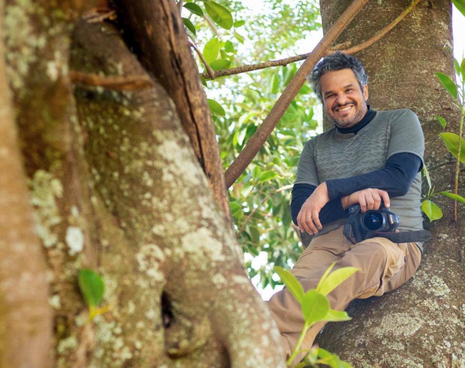Cristiano Xavier sorri, sentado no galho de uma grande árvore