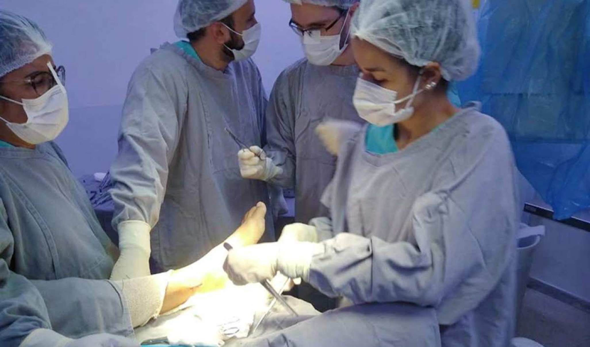 MÃ©dicos dentro de uma sala no Hospital realizando uma cirurgia. Os mÃ©dicos vestem roupas azuis e mÃ¡scaras.