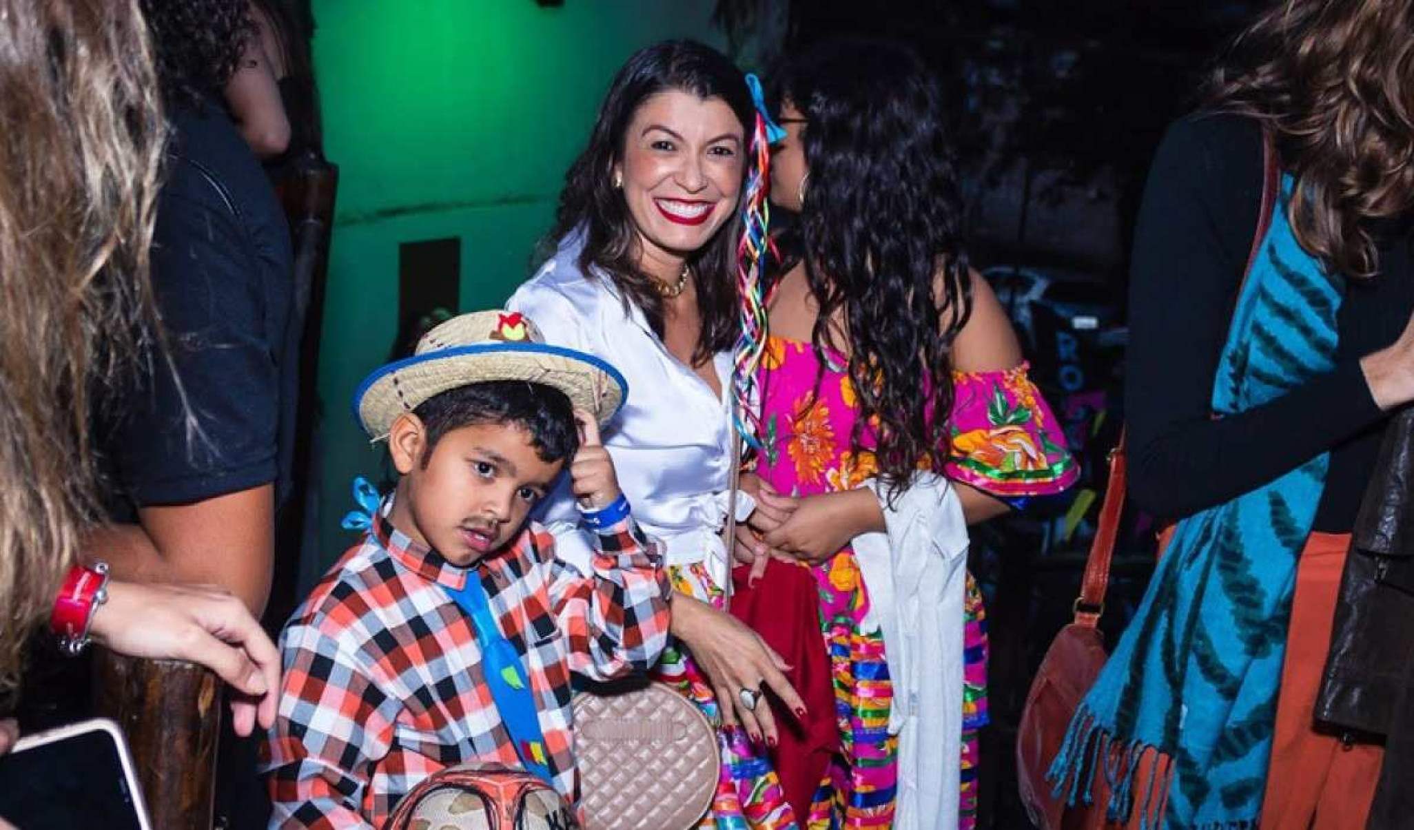 Mulher ao lado de duas crianças, uma menina e um menino. Todos vestidos com roupa de festa junina.