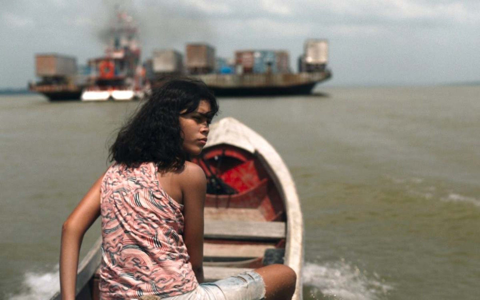 Atriz Jamilli Correa olha para trás, sentada em barco que navega pelo rio no filme Manas