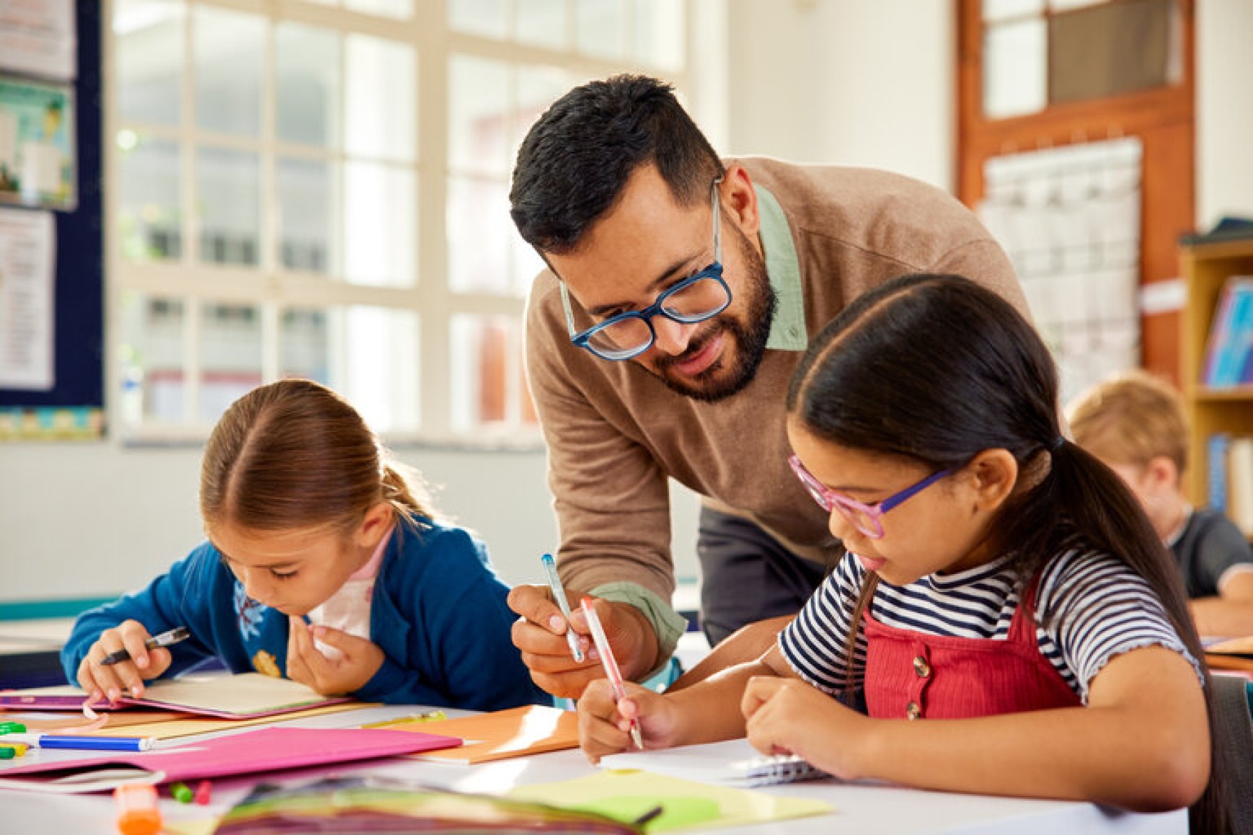  Male teacher in classroom wearing eyeglasses and helping school kids at desk. Little girls in classroom asking for help in solving sums to tutor. Male teacher helping elementary students in understanding concept while studying.
     -  (crédito:  francescoridolfi.com)