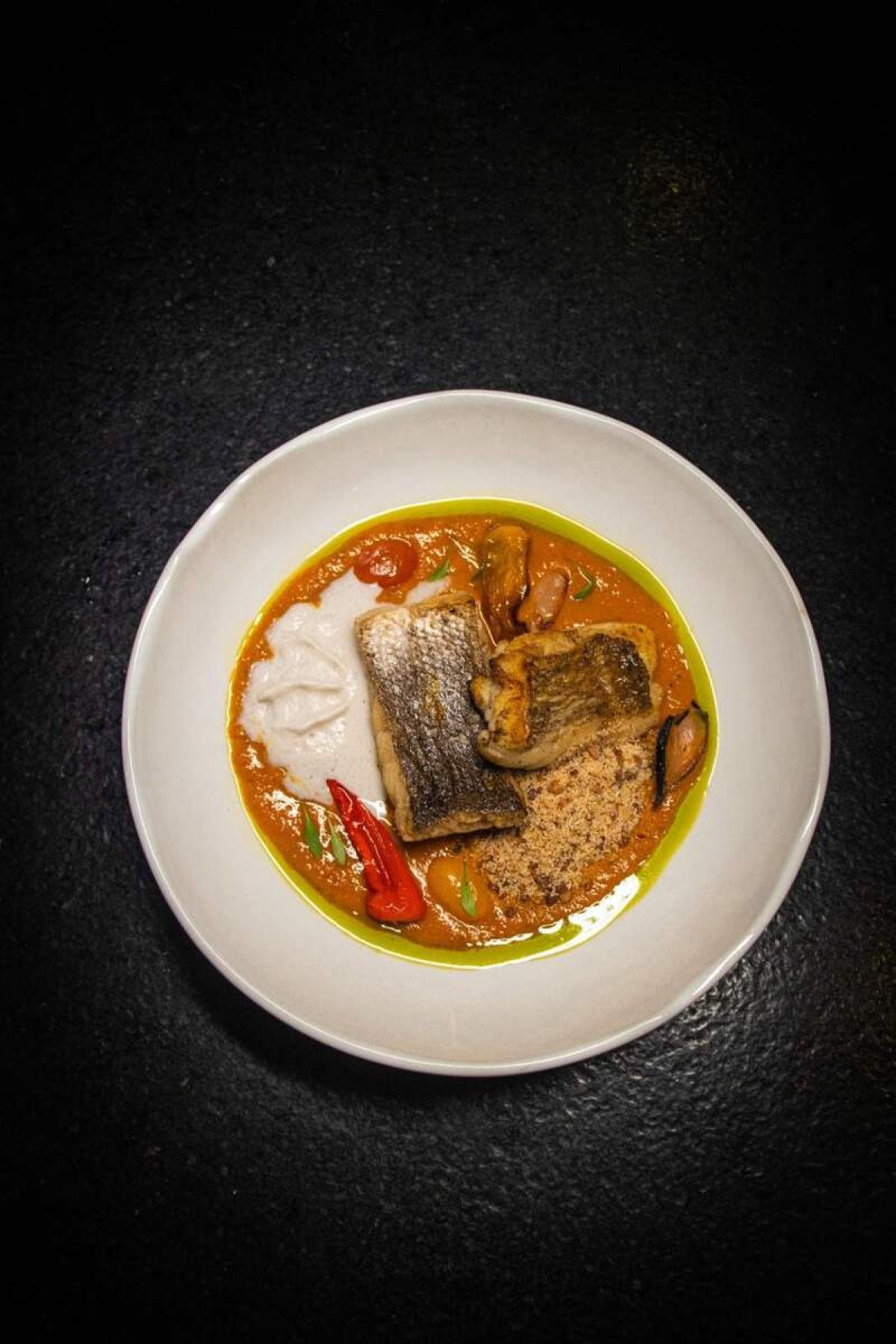 Na foto moqueca de peixe branco. Inspirado pela exuberância tropical e pela rica herança culinária do Brasil, este prato celebra a moqueca, um prato tradicional da cozinha baiana. A moqueca de peixe branco, cozida em um molho de leite de coco, dendê e coentro, é uma homenagem aos sabores vibrantes da Bahia. A espuma de coco queimado adiciona uma textura leve e um sabor tostado, enquanto a farofa de baru, uma castanha típica do cerrado brasileiro, oferece um contraste crocante. Acompanha arroz. Na foto moqueca de peixe branco. Inspirado pela exuberância tropical e pela rica herança culinária do Brasil, este prato celebra a moqueca, um prato tradicional da cozinha baiana. A moqueca de peixe branco, cozida em um molho de leite de coco, dendê e coentro, é uma homenagem aos sabores vibrantes da Bahia. A espuma de coco queimado adiciona uma textura leve e um sabor tostado, enquanto a farofa de baru, uma castanha típica do cerrado brasileiro, oferece um contraste crocante. Acompanha arroz.