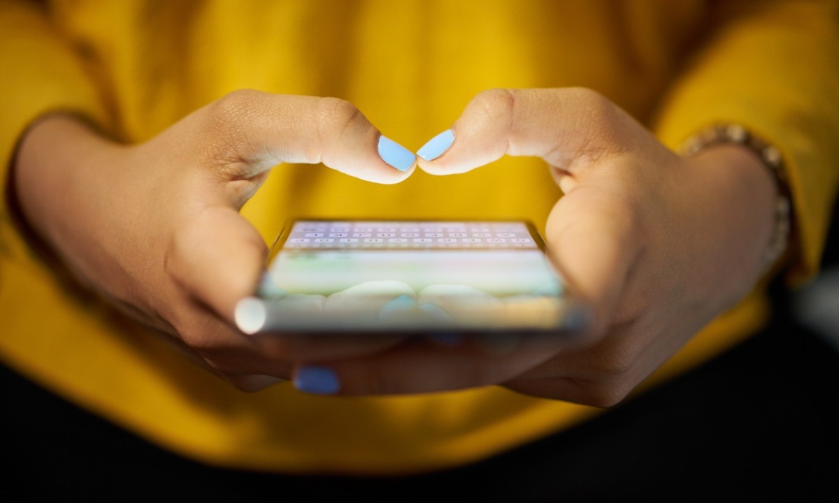  Young woman using cell phone to send text message on social network at night. Closeup of hands with computer laptop in background
     -  (crédito:  Getty Images/iStockphoto)