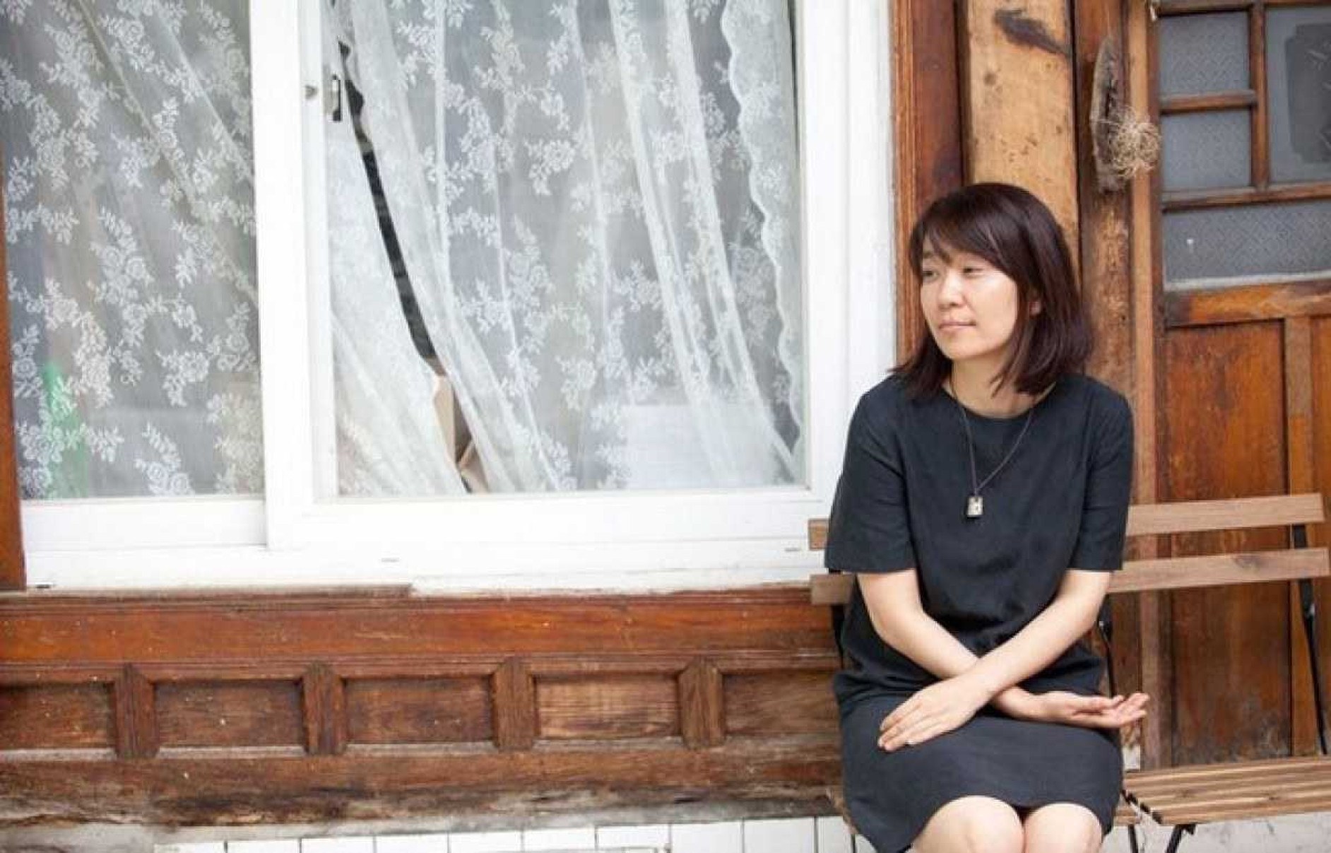Escritora sul-coreana Han Kang está sentada em um banco à frente de casa com janela onde se vê cortina branca