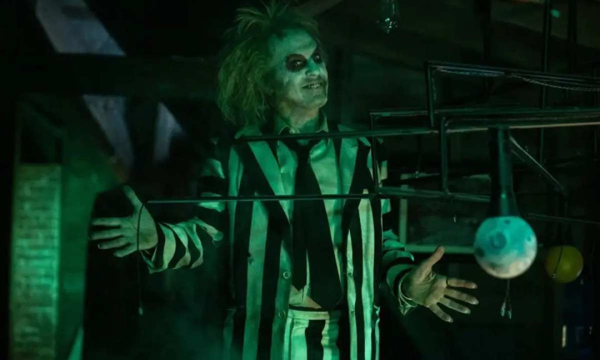 Beetlejuice, interpretado por Michael Keaton, homem branco com maquiagem preta e branca na cara. Ele está com o cabelo espetado e este terno listrado também peto e branco. 