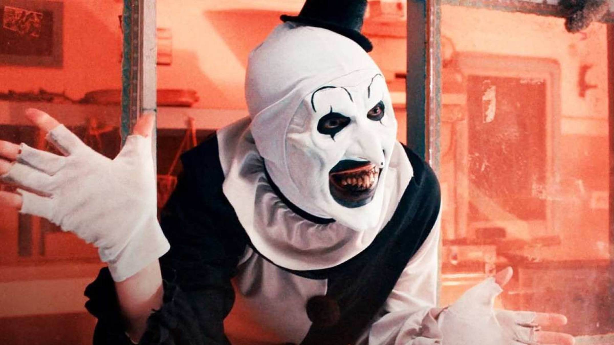 Terrifier, palhaço que utiliza maquiagens branca e preta. Sua roupa é típica de palhaço e é nas mesmas cores de sua maquiagem. Na foto ele sorri e está com ambas as mãos abertas.