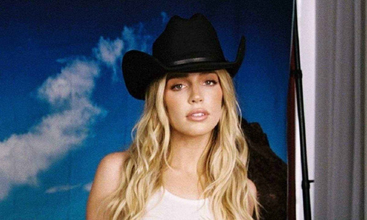 Alex Cooper, mulher branca com cabelo comprido, ondulado e loio. Ela usa uma regata branca e chapéu de cowboy preto. 