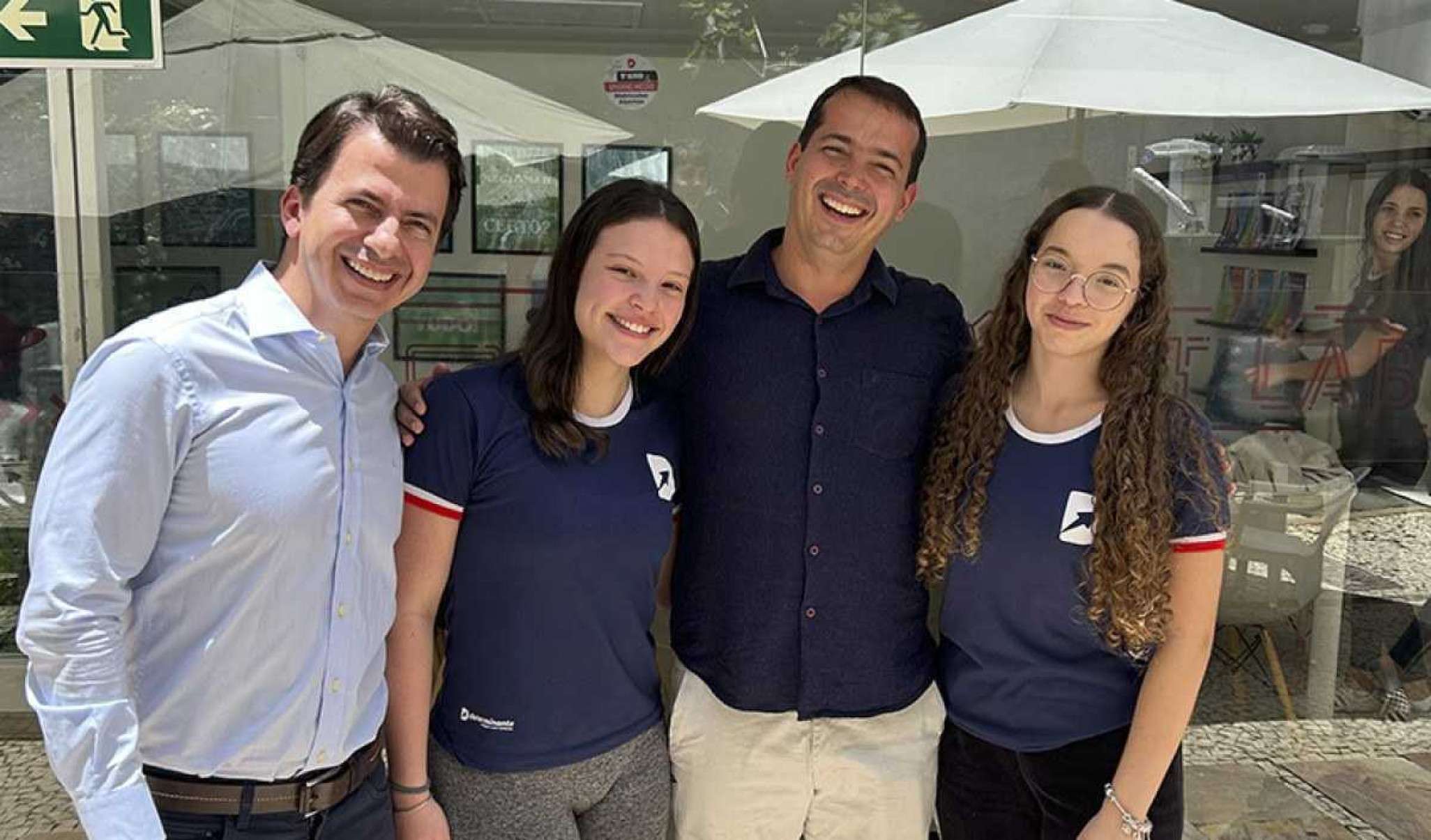 Dois adultos ao lados de duas jovens a frente de uma sala de estudos de vidro.