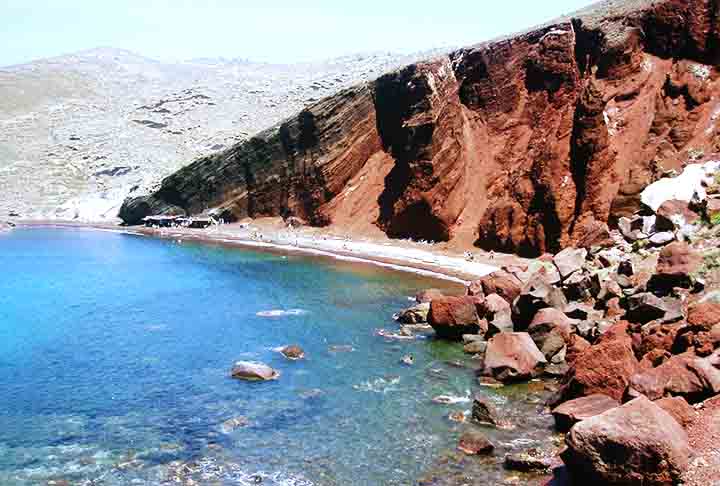 Praia de Red Beach – Santorini, Grécia - Cercada por penhascos de rocha vulcânica vermelha, sua areia avermelhada e pedras cria uma aparência marcante e única na ilha grega.