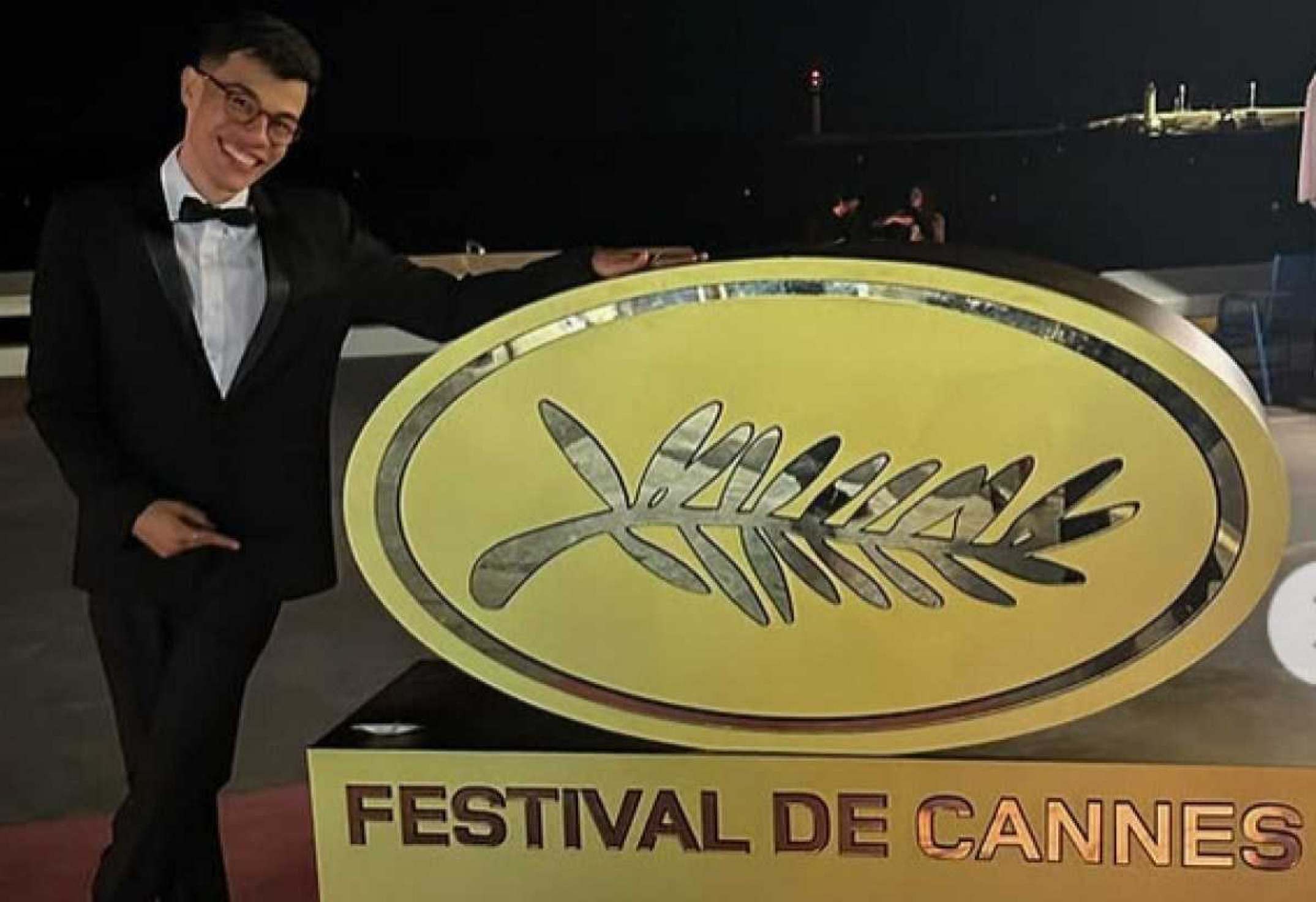 Ator João Pedro Mariano sorri e aponta para placa indicativa do Festival de Cannes, na França, em maio de 2024