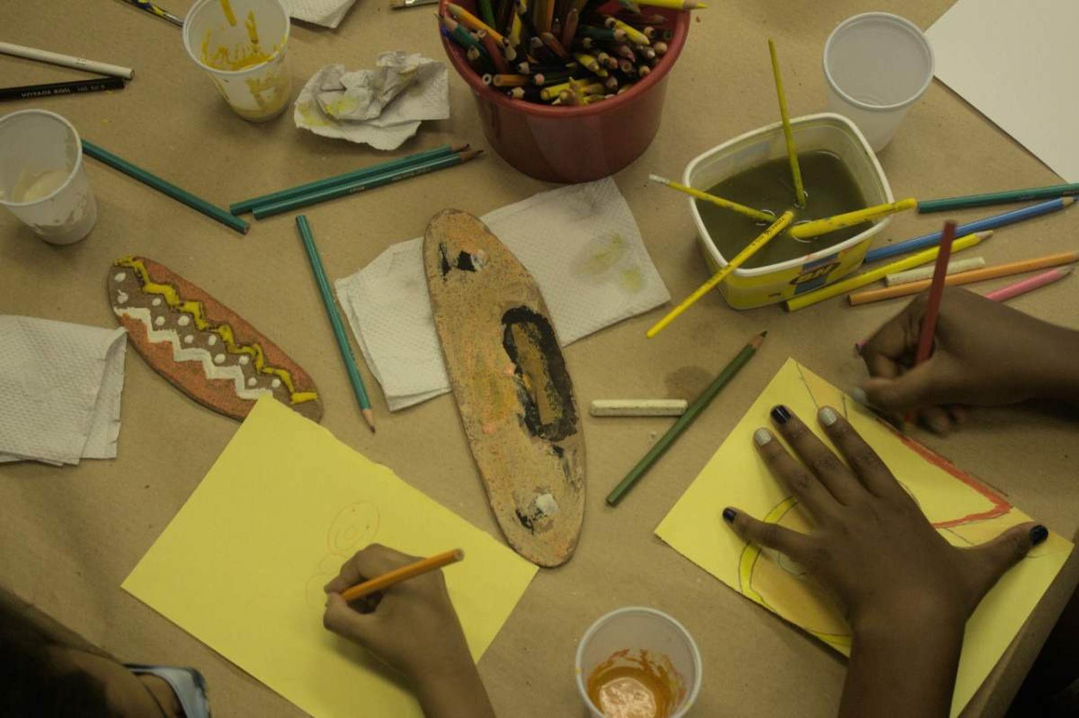 Foto mostra mãos de adultos e crianças recortando e desenhando máscaras em oficina da UFMG 