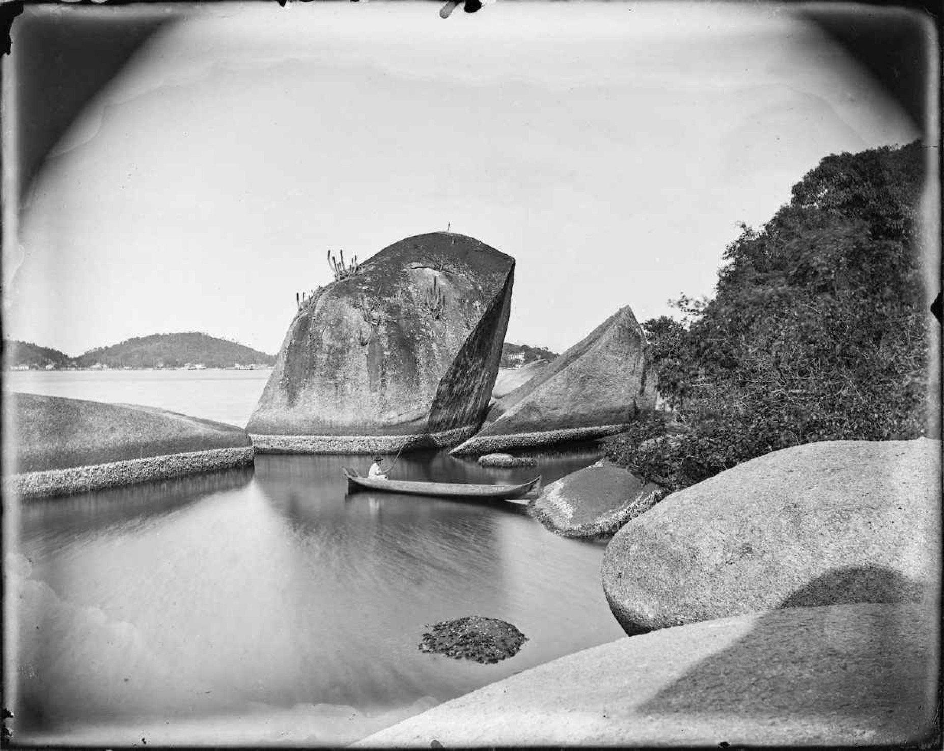 Fotografia feita por Marc Ferrez mostra o mar do Rio de Janeiro, entre pedras, com um pequeno barco ao centro 