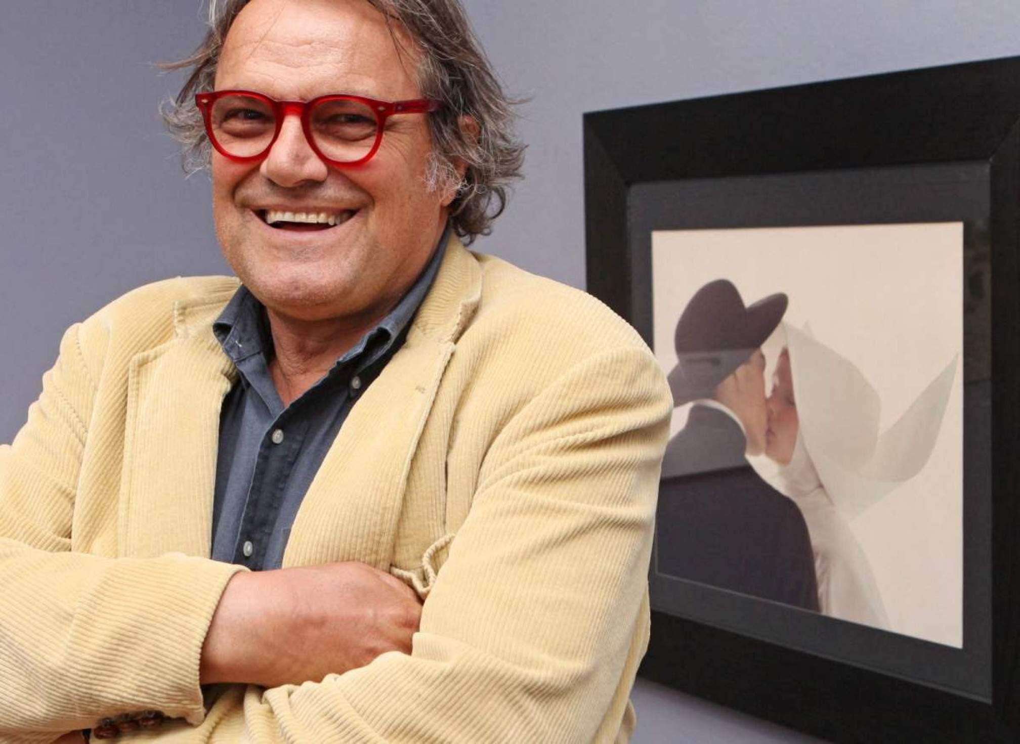 Fotógrafo Oliviero Toscani sorri, de braços cruzados, à frente de tela que exibe campanha publicitária feita por ele para a grife Benetton