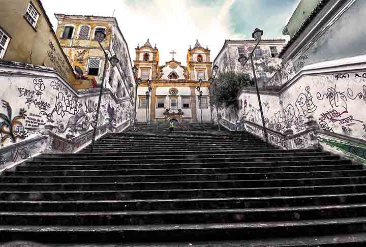 Escadaria da Igreja do Santíssimo Sacramento da Rua do Passo - Salvador (BA) - A escadaria conecta o bairro do Santo Antônio Além do Carmo ao Pelourinho e é famosa por sua arquitetura colonial e seu charme histórico. Composta por mais de 50 degraus de pedra, é cercada por casas coloridas e possui grande relevância no contexto cultural e religioso. Cenário de filmes e novelas, a escadaria evoca a essência do centro histórico de Salvador.