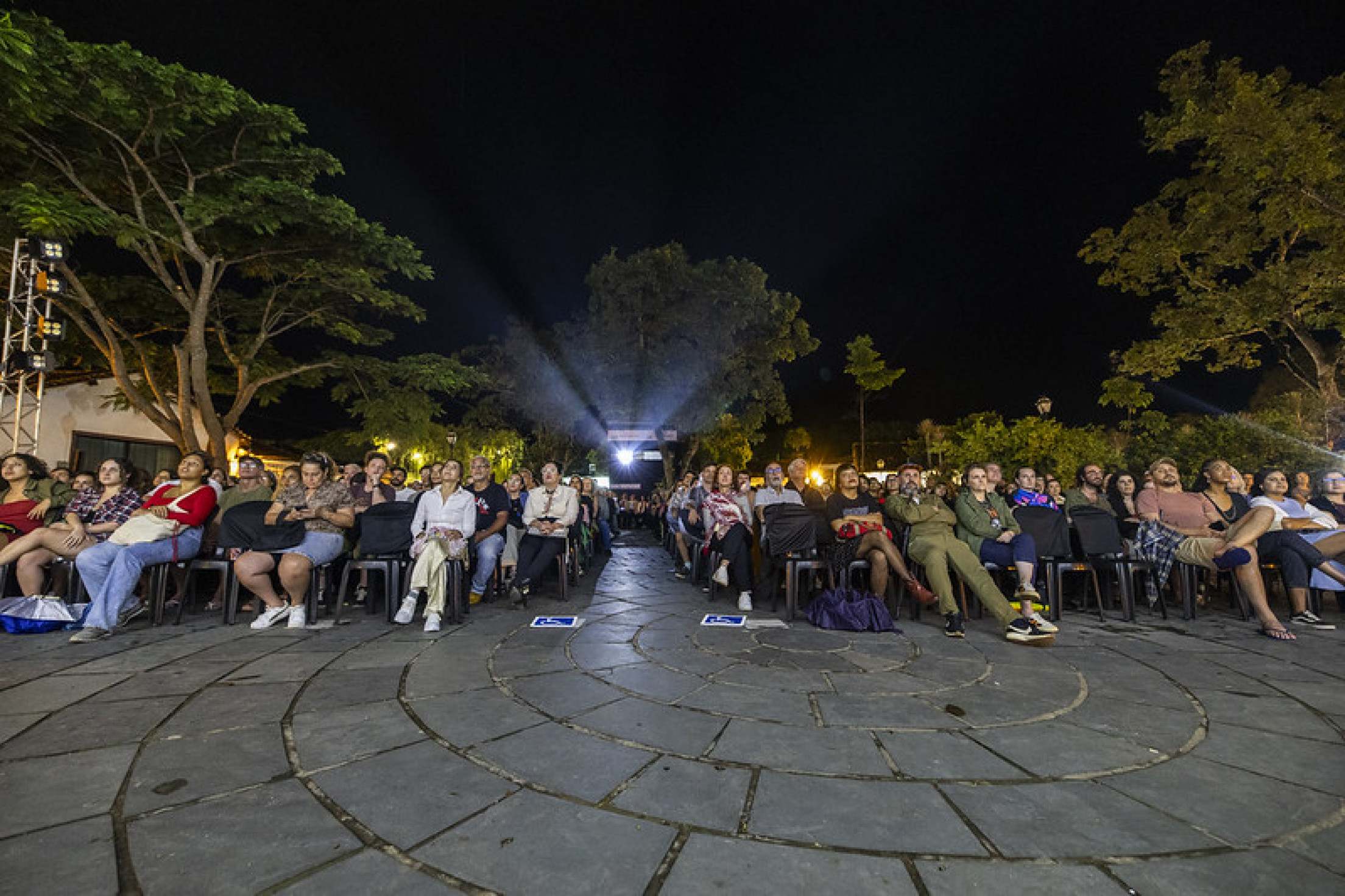 Pessoas estão assistindo a filme na praça, ao ar livre, em Tiradentes. Ao fundo, vê-se a luz do projetor