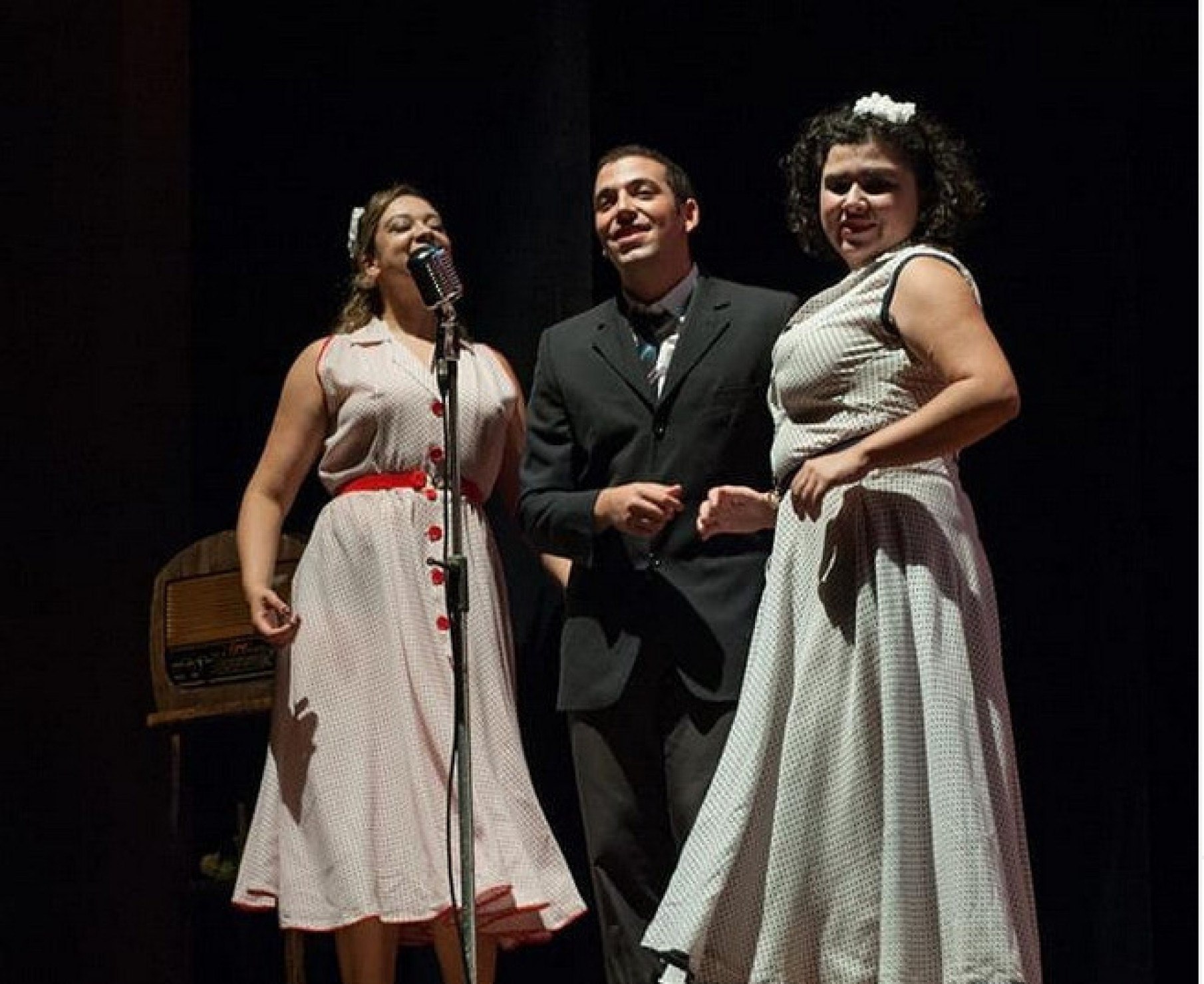 Duas atrizes e um ator interpretam cantores, em frente ao microfone, no musical Nas ondas do rádio, em BH