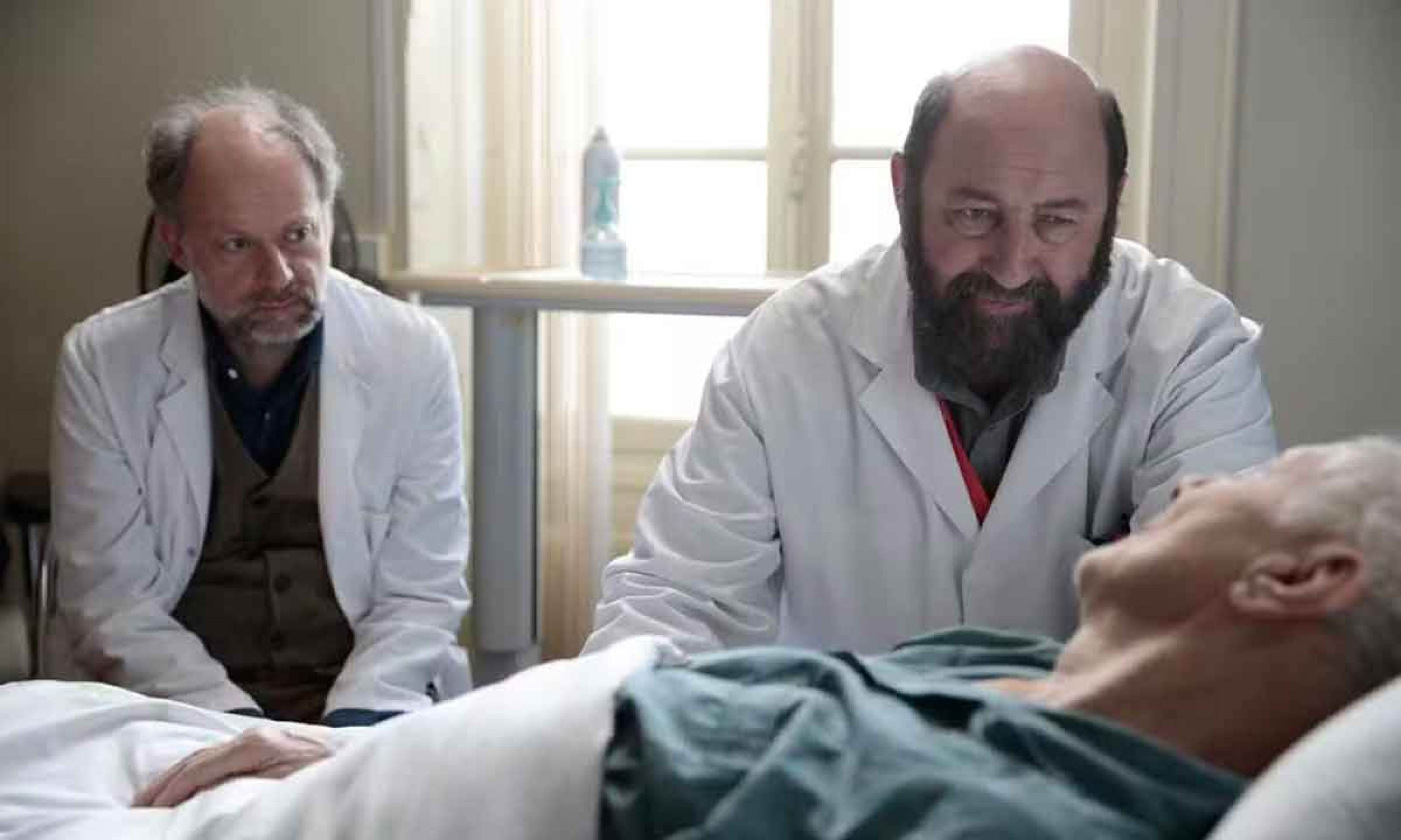 Em cena do filme Le dernier souffle, os atores Denis Podalydès e Kad Merad estão sentados lado a lado, em frente a paciente deitado em cama do hospital