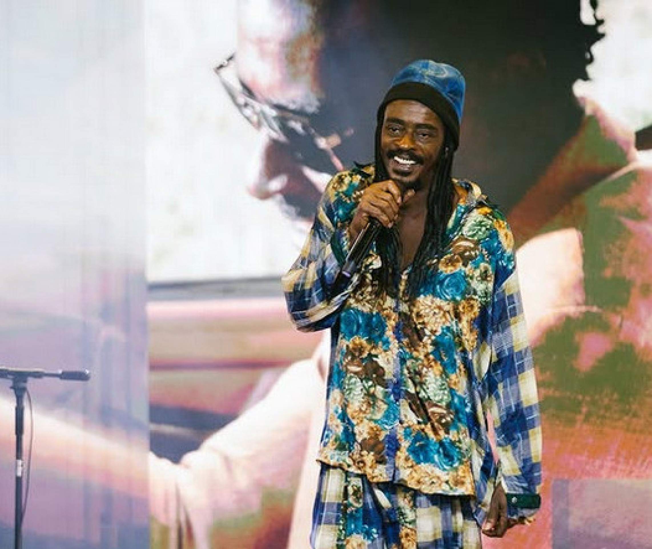 O cantor e compositor Seu Jorge, vestindo roupa colorida, canta no palco, à frente de telão com sua própria imagem