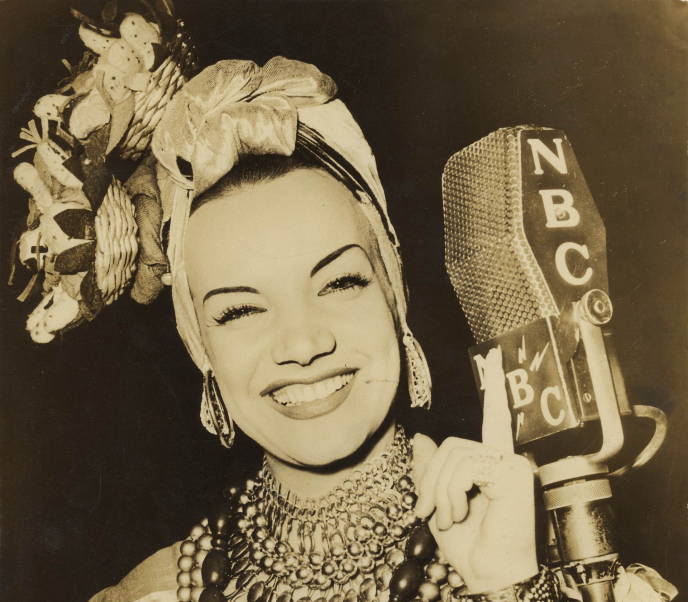  Carmen Miranda usando turbante estilizado de baiana ri para a câmera em frente a microfone antigo, em foto de 1040