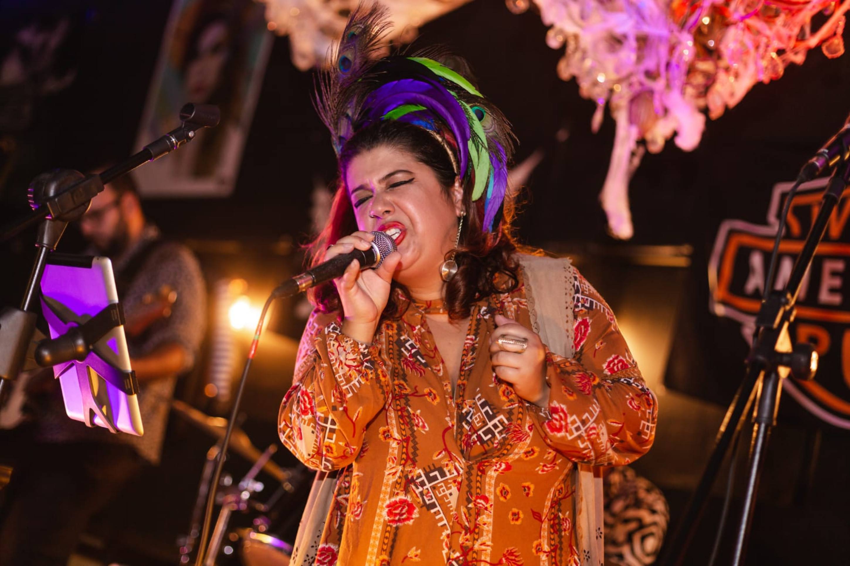 Cantar o repertório de Janis Joplin exige de Luiza Frad's muitos ensaios e aulas de técnica vocal Vestindo figurino laranja, Luiza Frad's canta ao microfone
