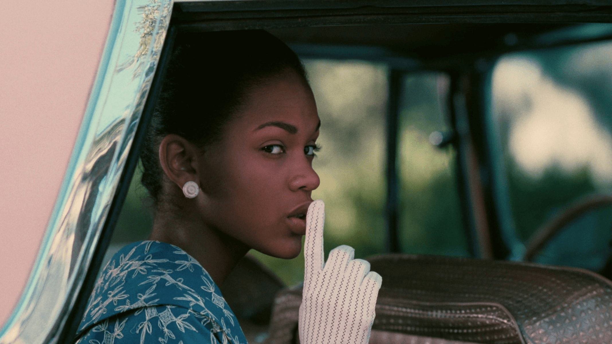 Atriz olha da janela de carro e faz o sinal de silêncio com a mão nos lábios, em cena do filme Amores divididos 