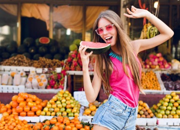 Garota sorridente, com cabelos longos e claros, faz pose em banca de mercado, com frutas tropicais ao fundo
     -  (crédito:  Vadim Fedorchenko/freepik)
