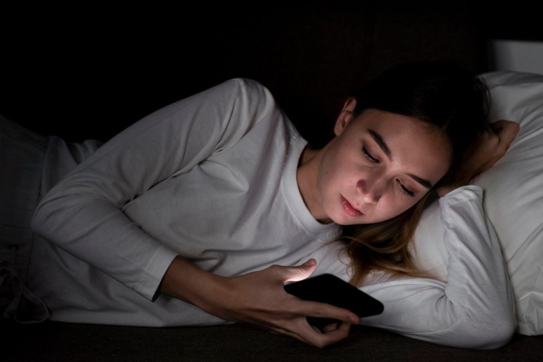Usar o celular antes de dormir pode prejudicar intensamente sua noite de sono Pessoa com celular na mão no quarto escuto, preparada para dormir