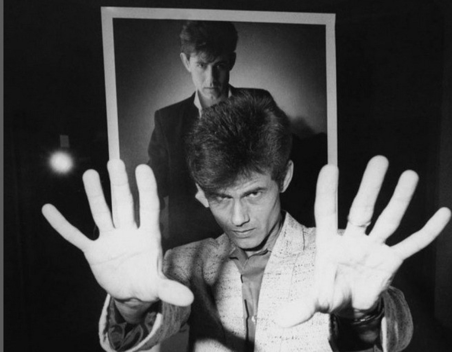 Cantor Ritchie com as mãos espalmadas em frente ao rosto, nos anos 1980, tendo atrás de si uma foto de divulgação de seus discos