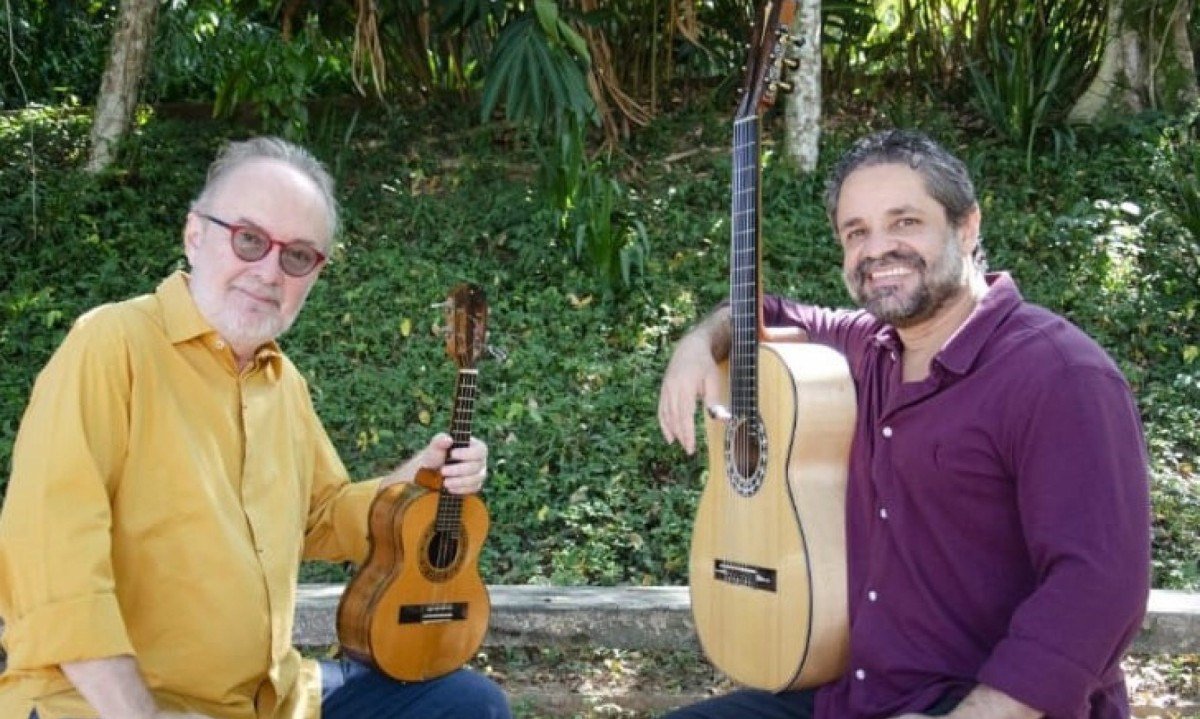  Músicos Henrique Cazes e Rogério Caetano seguram instrumentos musicais, estão frente a frente e sorriem para a câmera