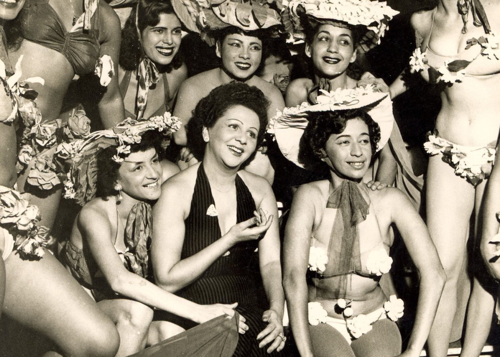Nos anos 1950, cantora Dalva de Oliveira, de vestido preto, está rodeada de vedetes de biquini e sorri para a câmera