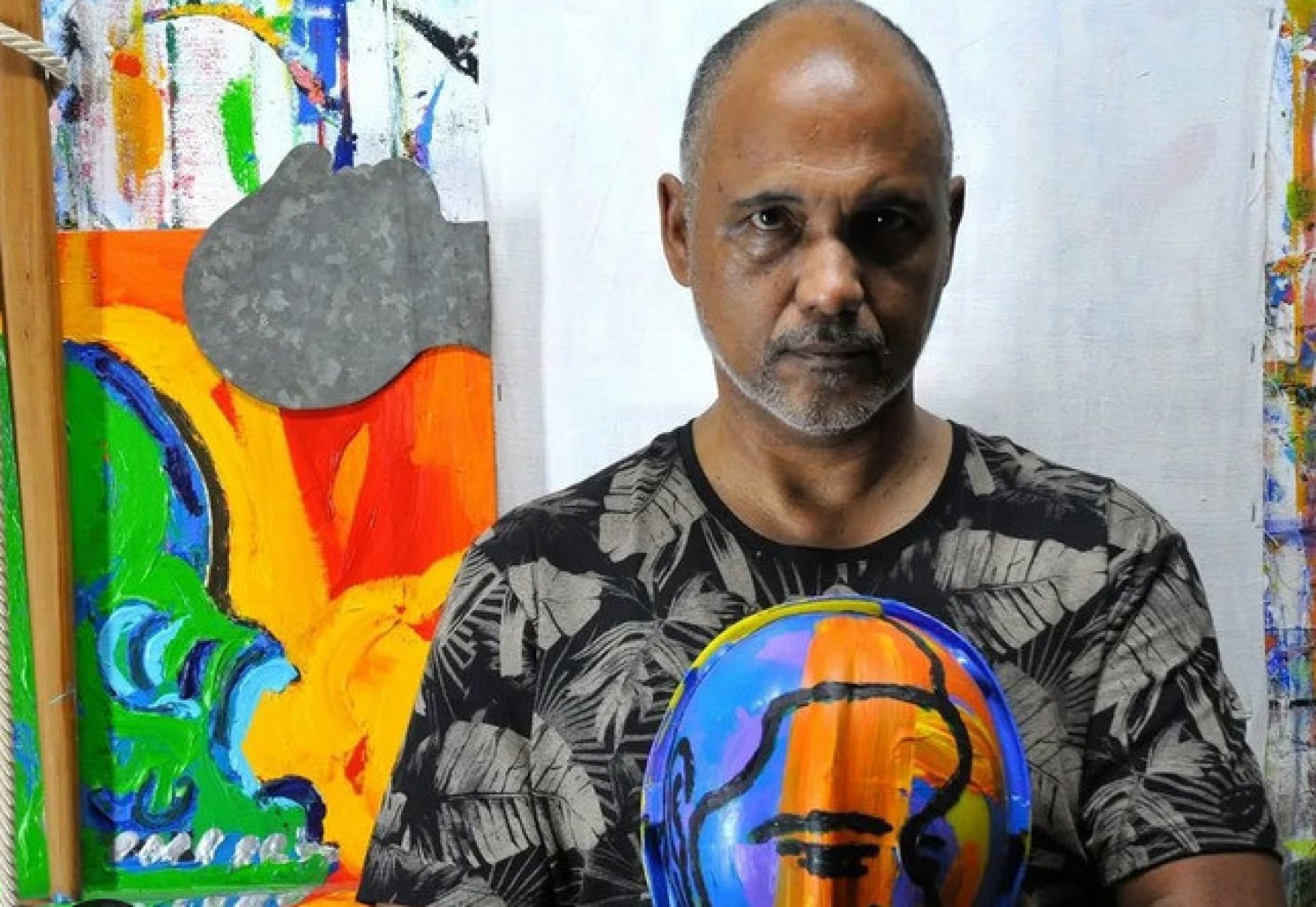 O artista plástico mineiro mineiro Antonio Sérgio Moreira, à frente de um quadro colorido, olha para a câmera 