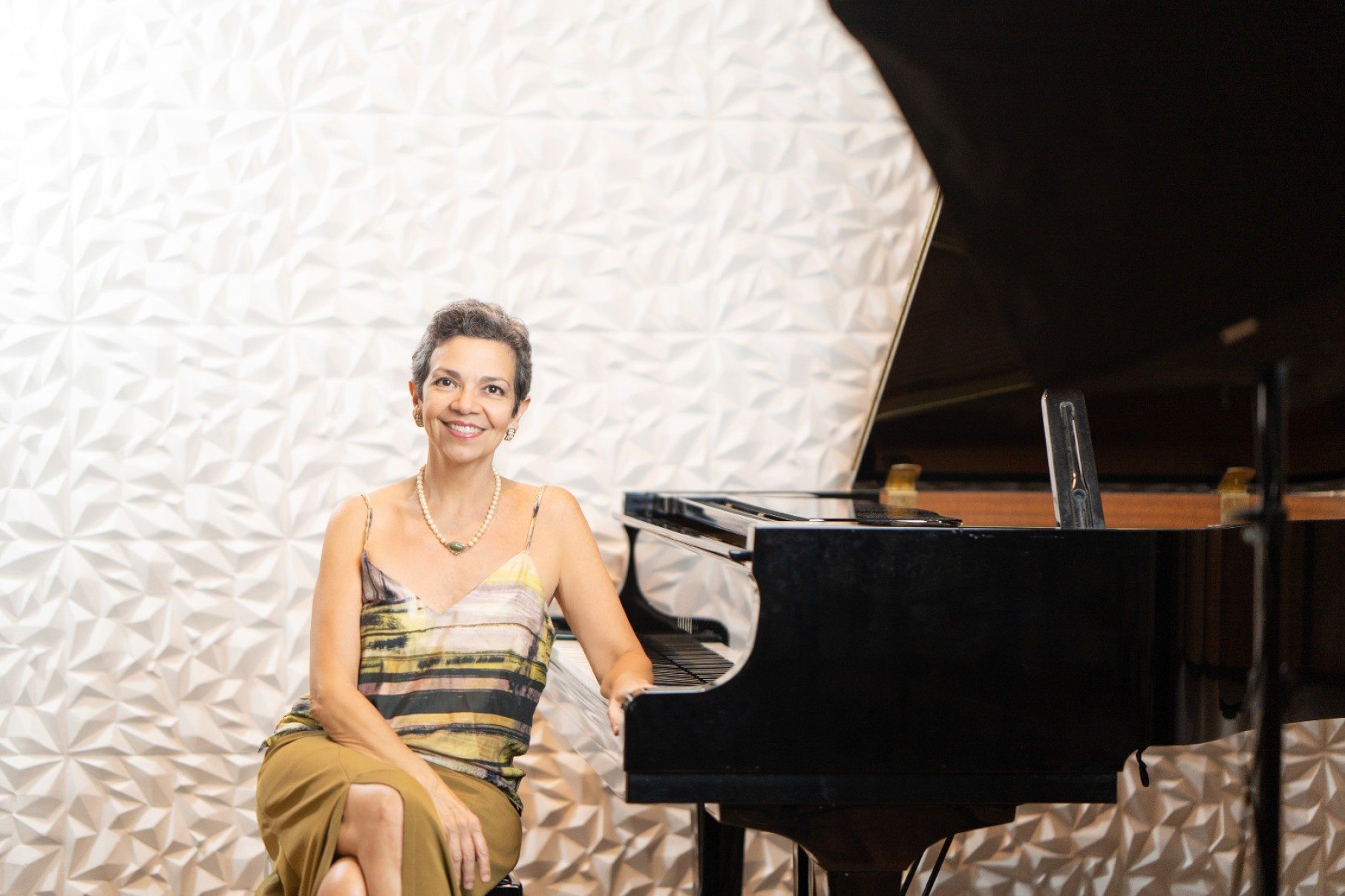 Pianista Maria Tereza Madeira está sentada ao lado do piano e sorri para a câmera