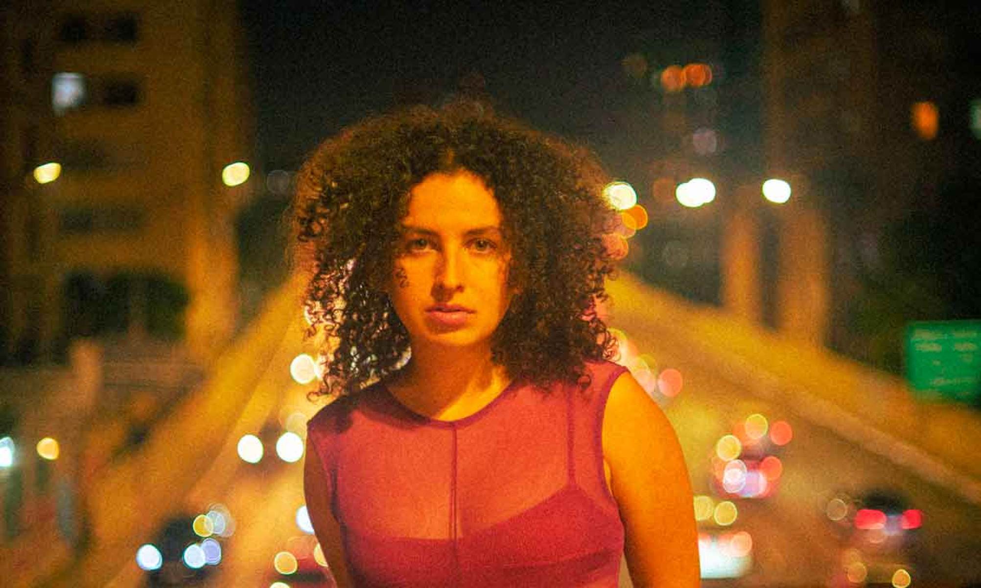 Além de composições próprias, Dora Morelenbaum cantará "Cucurrucucú Paloma" A compositora e cantora Dora Morelenbaum vestida de vermelho, com viaduto ao fundo