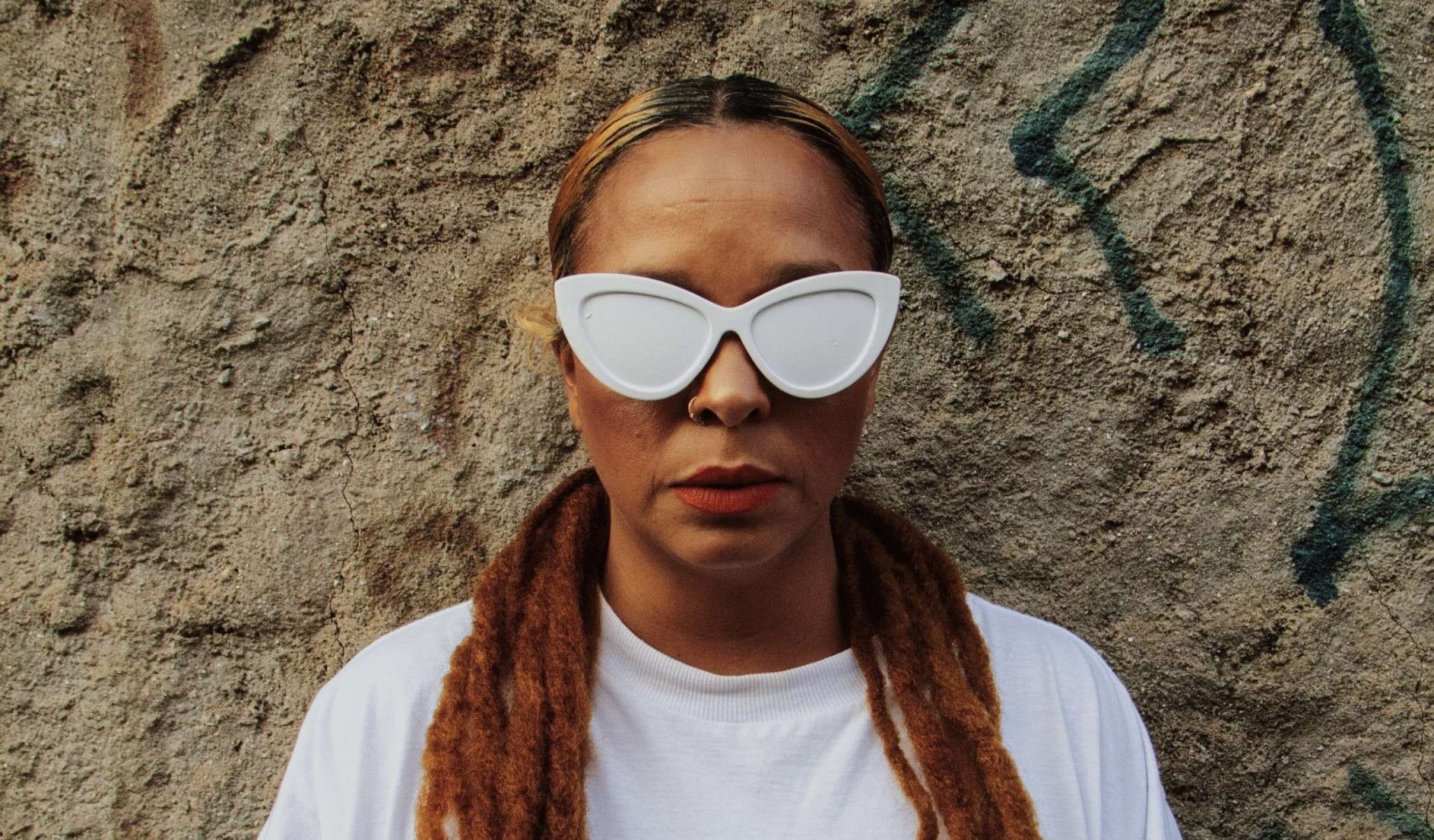 Anelis Assumpção, encostada na parede de chapisco,  usa camiseta branca e óculos com lentes pintadas de branco