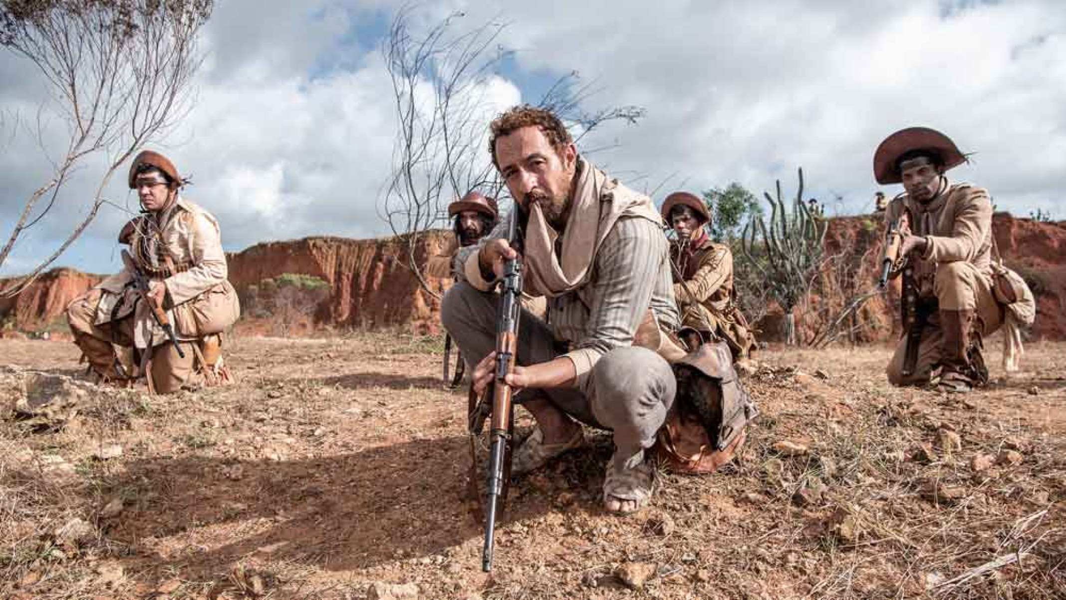 Ator Irandhir Santos está agachado, segurando espingarda, em meio a cangaceiros na novela Guerreiros do Sol 
