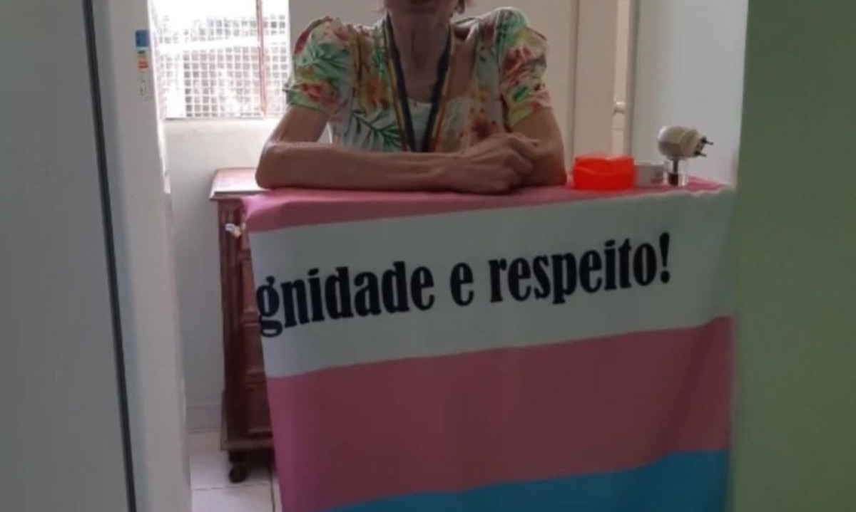 Sonia Sissy Kelly Lopes na bancada da cozinha, com a bandeira transgênero — em azul, rosa e branco — estampada com as palavras "dignidade e respeito"