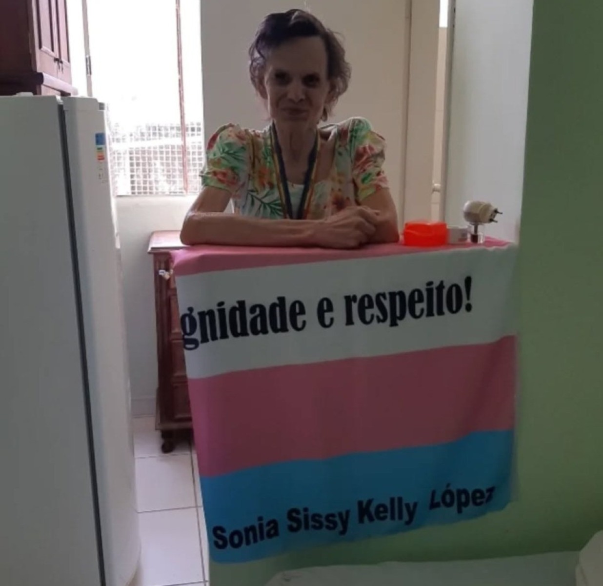 Sonia Sissy Kelly Lopes na bancada da cozinha, com a bandeira transgênero — em azul, rosa e branco — estampada com as palavras "dignidade e respeito"