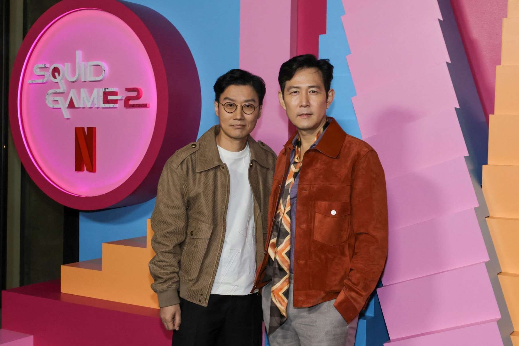O diretor Hwang Dong-hyuk, criador de 'Round 6', e o ator Lee Jung-jae, que interpreta o protagonista da atração, posam para a imprensa, tendo atrás deles painel colorido alusivo ao programa
