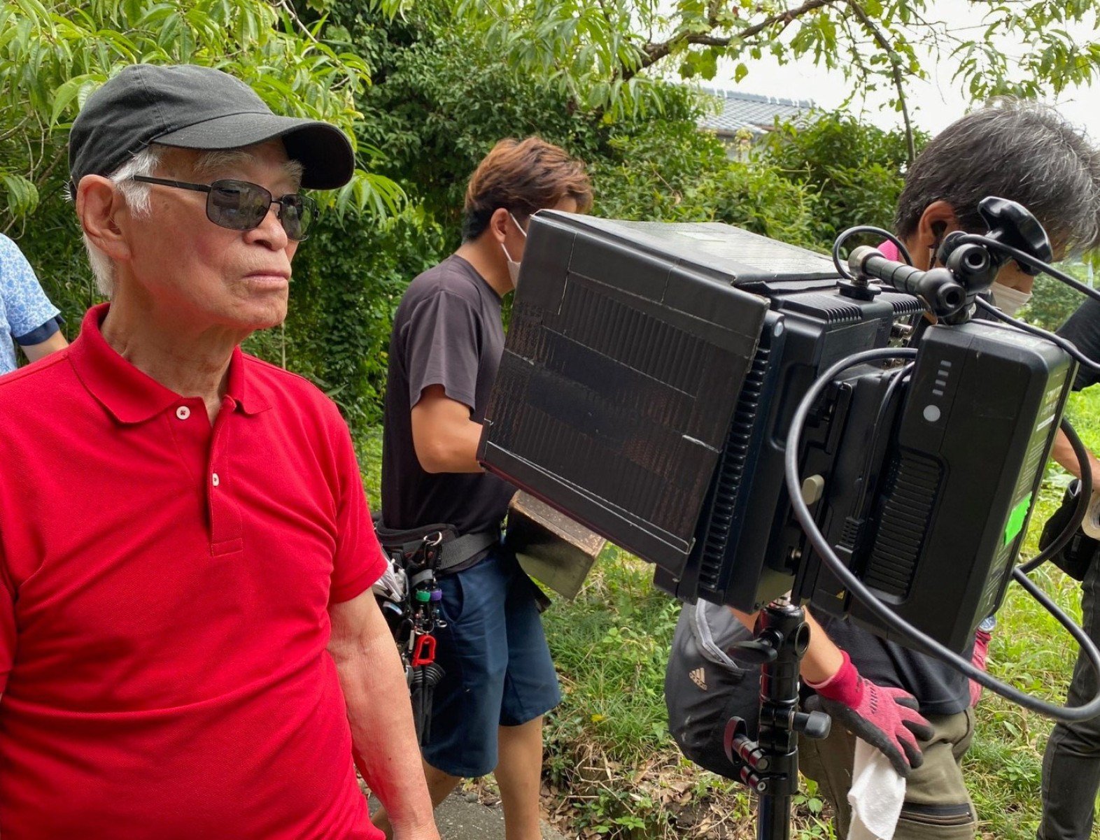 Diretor de cinema Masao Adachi, de boné verde e camisa vermelha, em frente à câmera de filmagem 