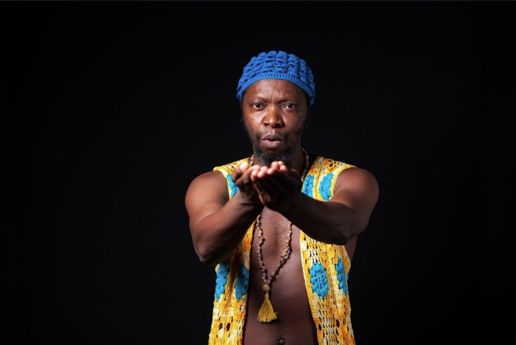 O cantor e compositor Sérgio Pererê usa turbante azul e estende os braços para a câmera