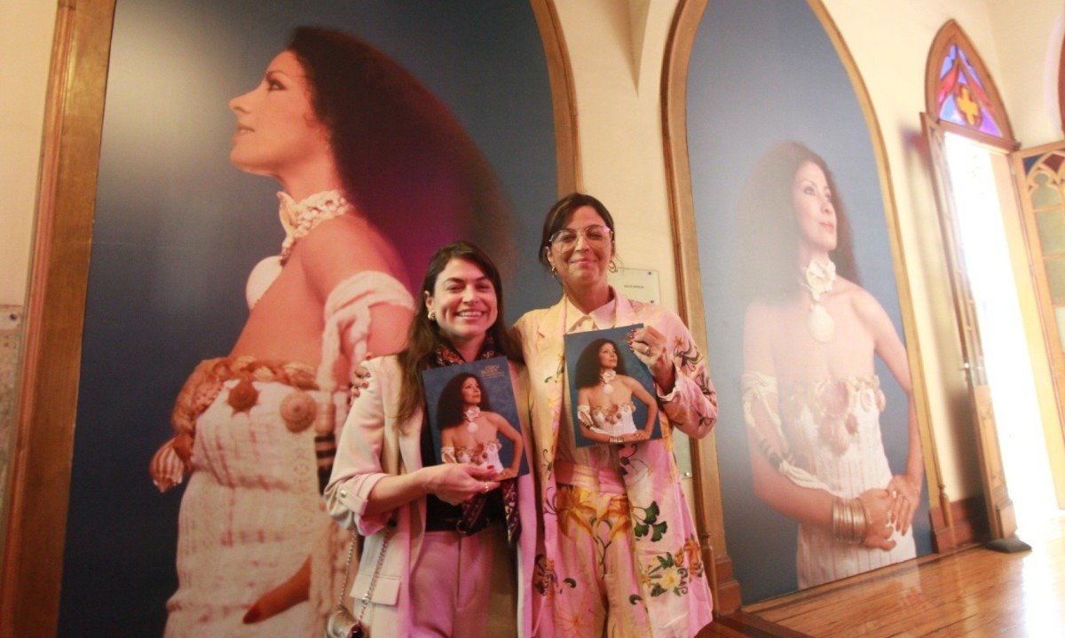 Carol Costa e Emanuelle Araújo no Museu da Moda de BH. As duas estão em frente a imensa foto da cantora Clara Nunes e seguram o programa da exposiçã, com o rosto de Clara na capa