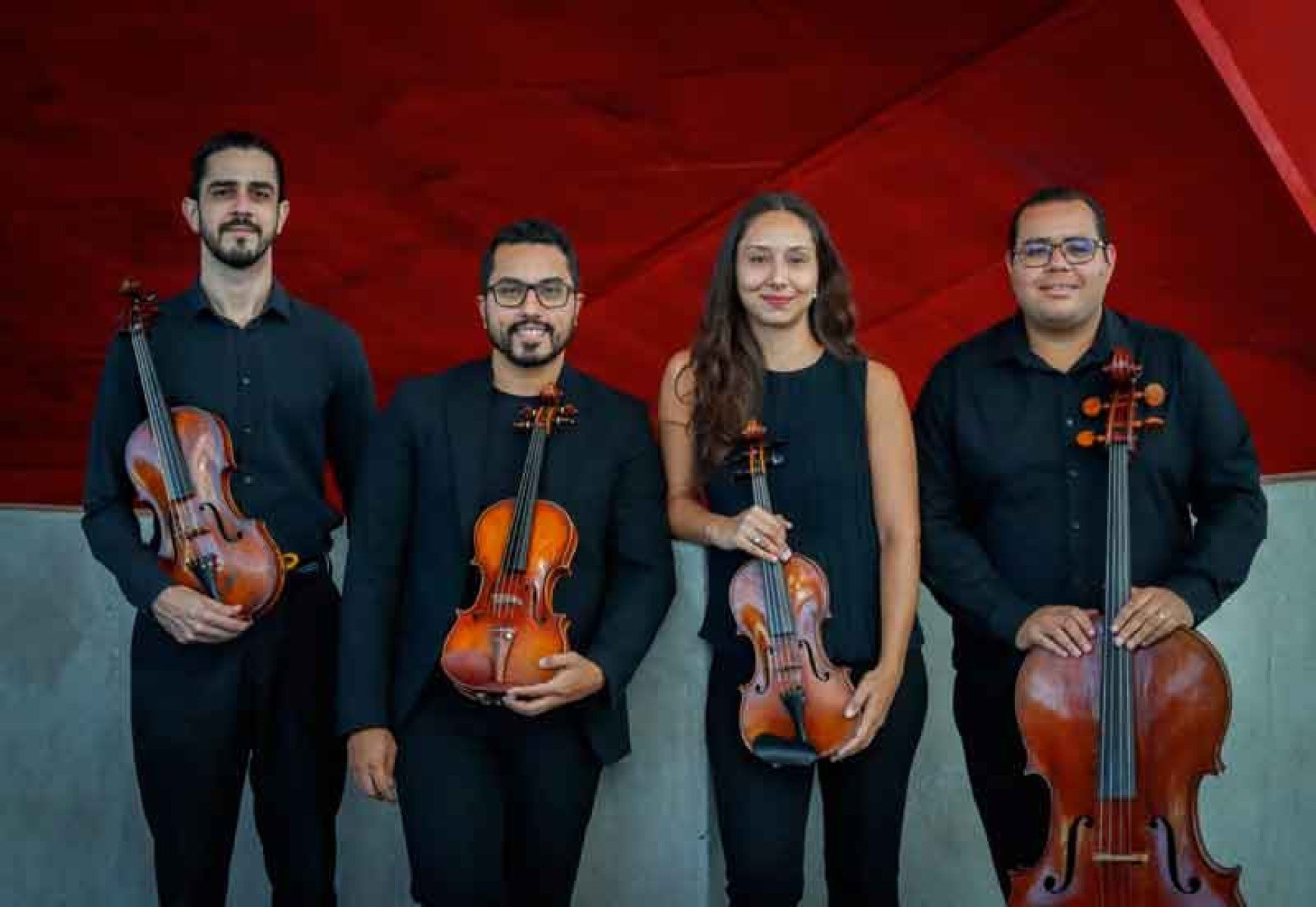Os integrantes do Quarteto Echinata, de pé, seguram seus instrumentos e sorriem, tendo ao fundo parede em tom de vermelho da Sala Minas Gerais