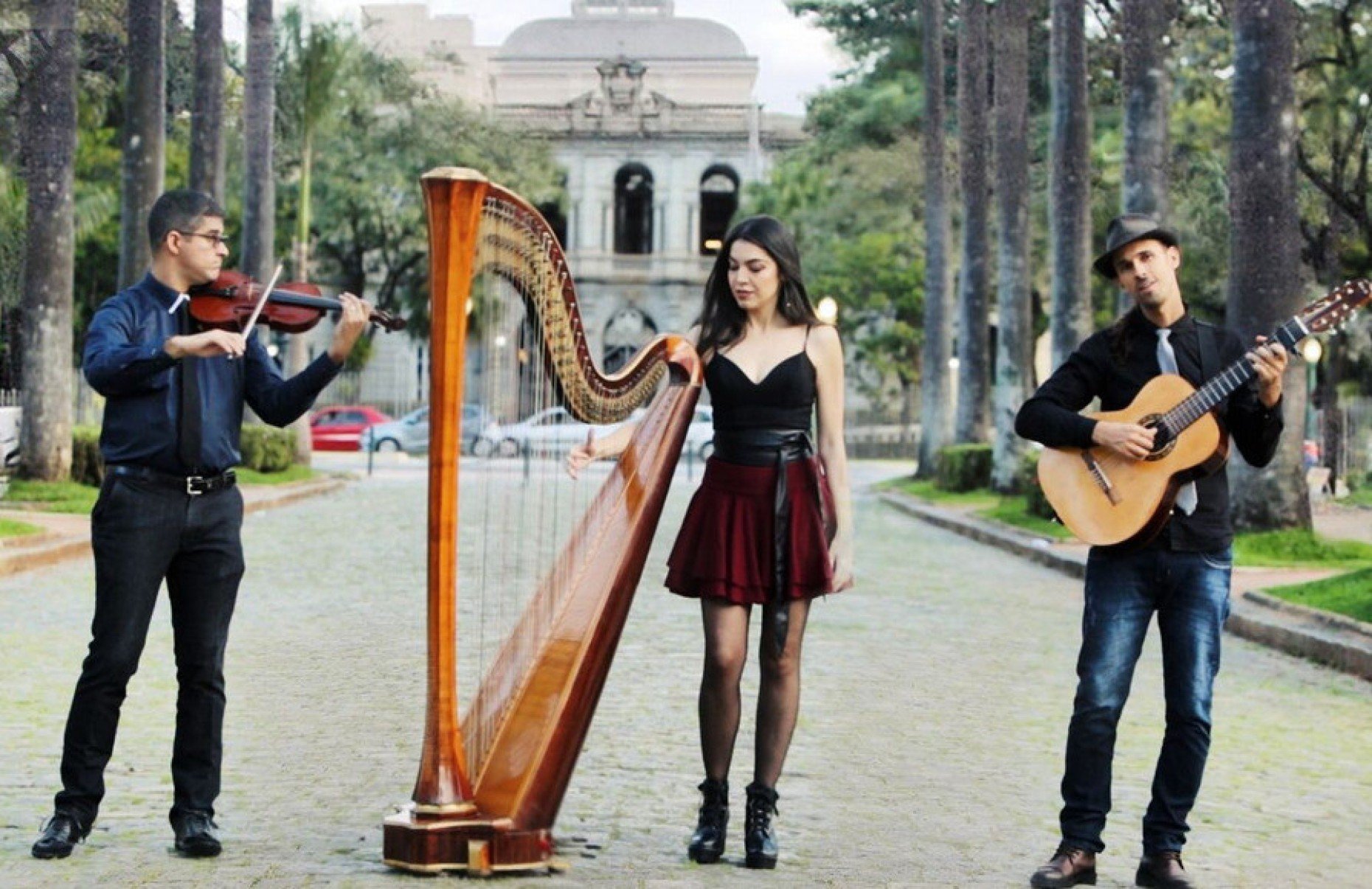 Músicos do Trio Amadeus, segurando violino, harpa e violão, posam na Praça da Liberdade, em BH, tendo o Palácio da Liberdade ao fundo