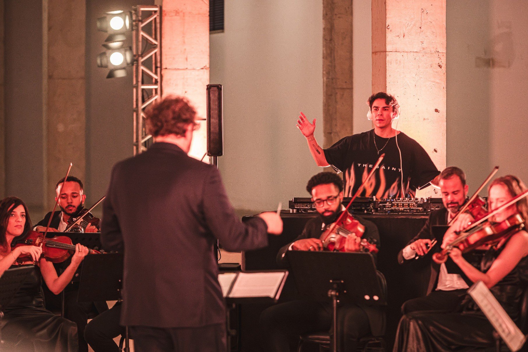 Maestro Rodrigo Toffolo, de costas para a câmera, rege orquestra tendo à frente dele o DJ Kvsh, com seus computadores