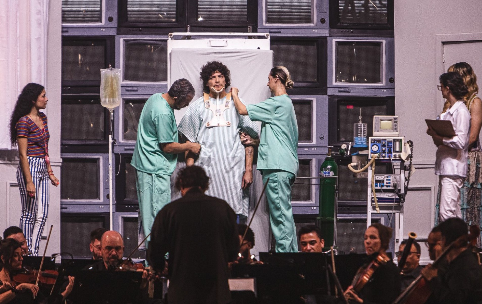 Personagem de ópera está na cama cercado de médicos. A cama está em pé, para poder ser vista pelo público